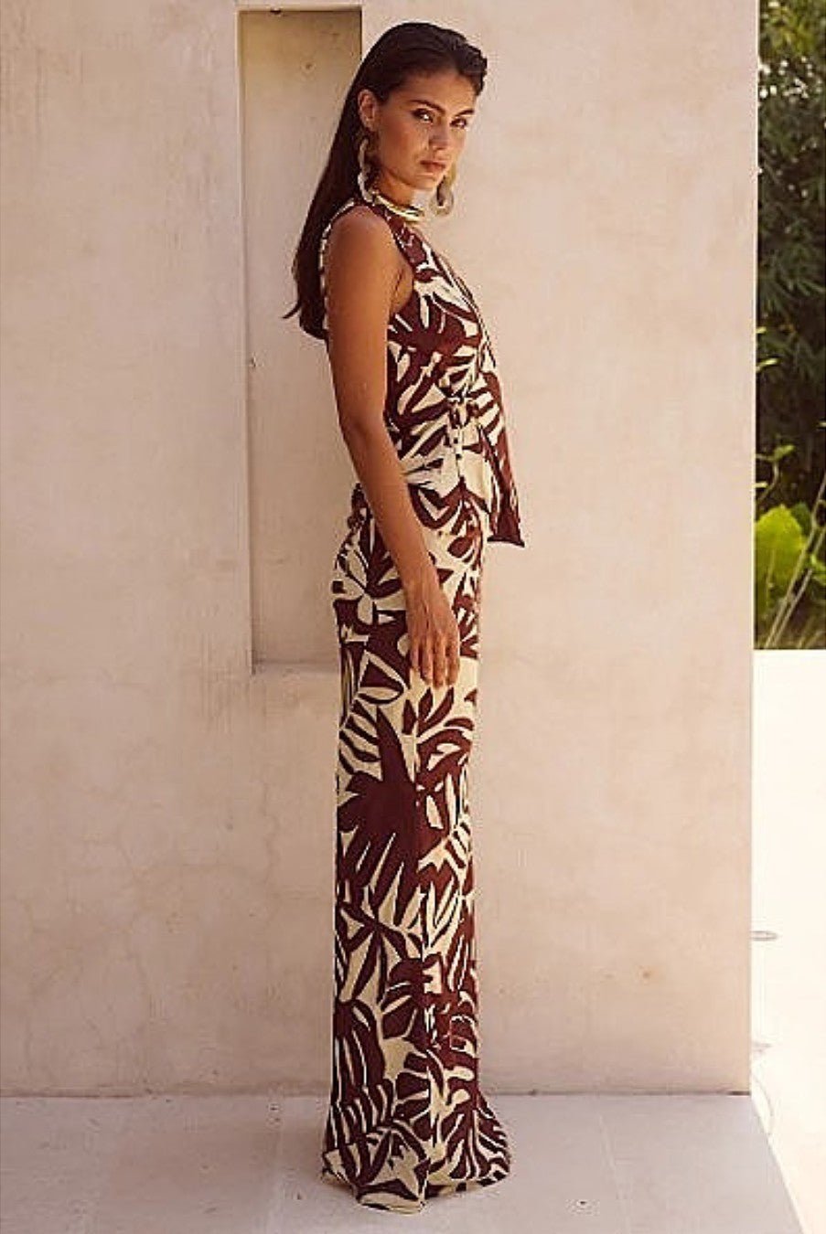 Model wearing brown printed maxi skirt set, standing against stone wall.