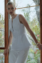 A model wearing the Mercer Sleeveless Top in white, standing by a window with trees outside.