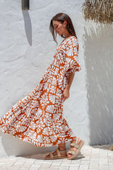 Woman wearing a long orange and white floral dress against a white wall.