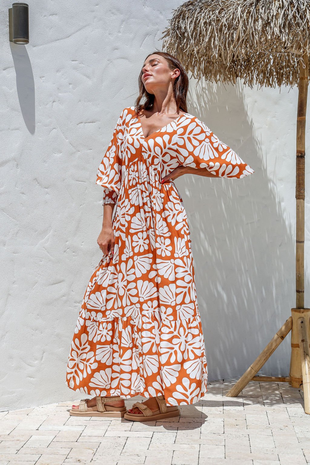 Woman wearing an orange and white floral dress against a white wall with a thatched roof.