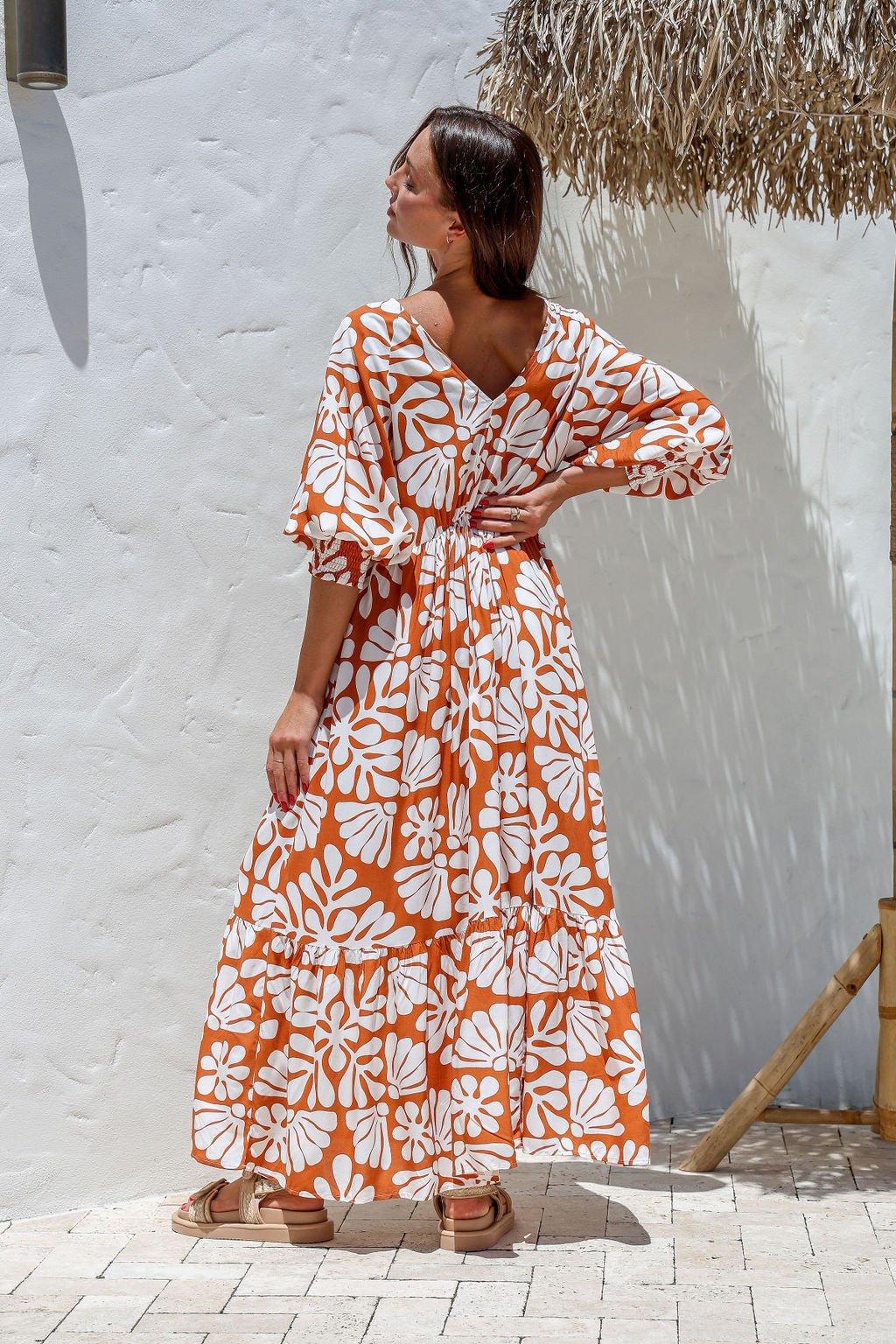 Woman wearing an orange and white floral dress standing against a white wall.