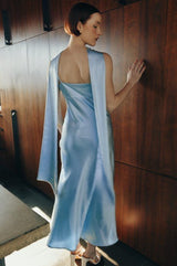 Woman in a light blue dress standing against a wooden wall.