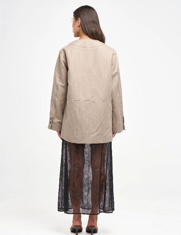 A woman shown from the back wearing a light beige linen-blend jacket with a black sheer lace maxi skirt, photographed against a plain white studio background.