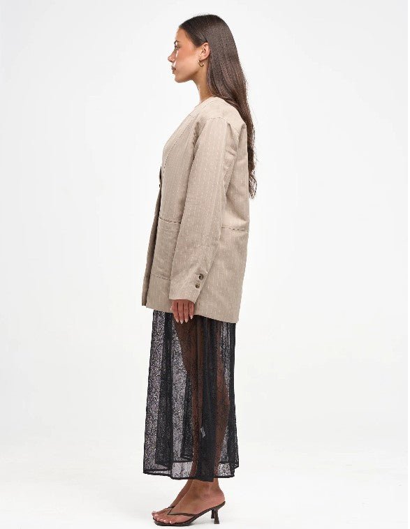A woman shown in profile wearing a light beige textured blazer over a sheer black maxi skirt, paired with brown heeled sandals, against a plain white studio background.