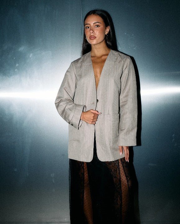 A woman modeling a light gray linen-textured blazer with black sheer side panels, paired with a black lace skirt, set against a dark gray studio background with a horizontal light streak.