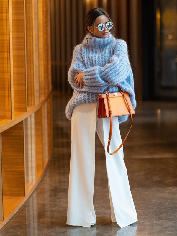 Woman in the blue Rainy Dayz Sweater with an orange shoulder bag and white pants in a modern indoor setting.