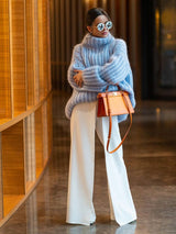 Woman in the blue Rainy Dayz Sweater with an orange shoulder bag and white pants in a modern indoor setting.