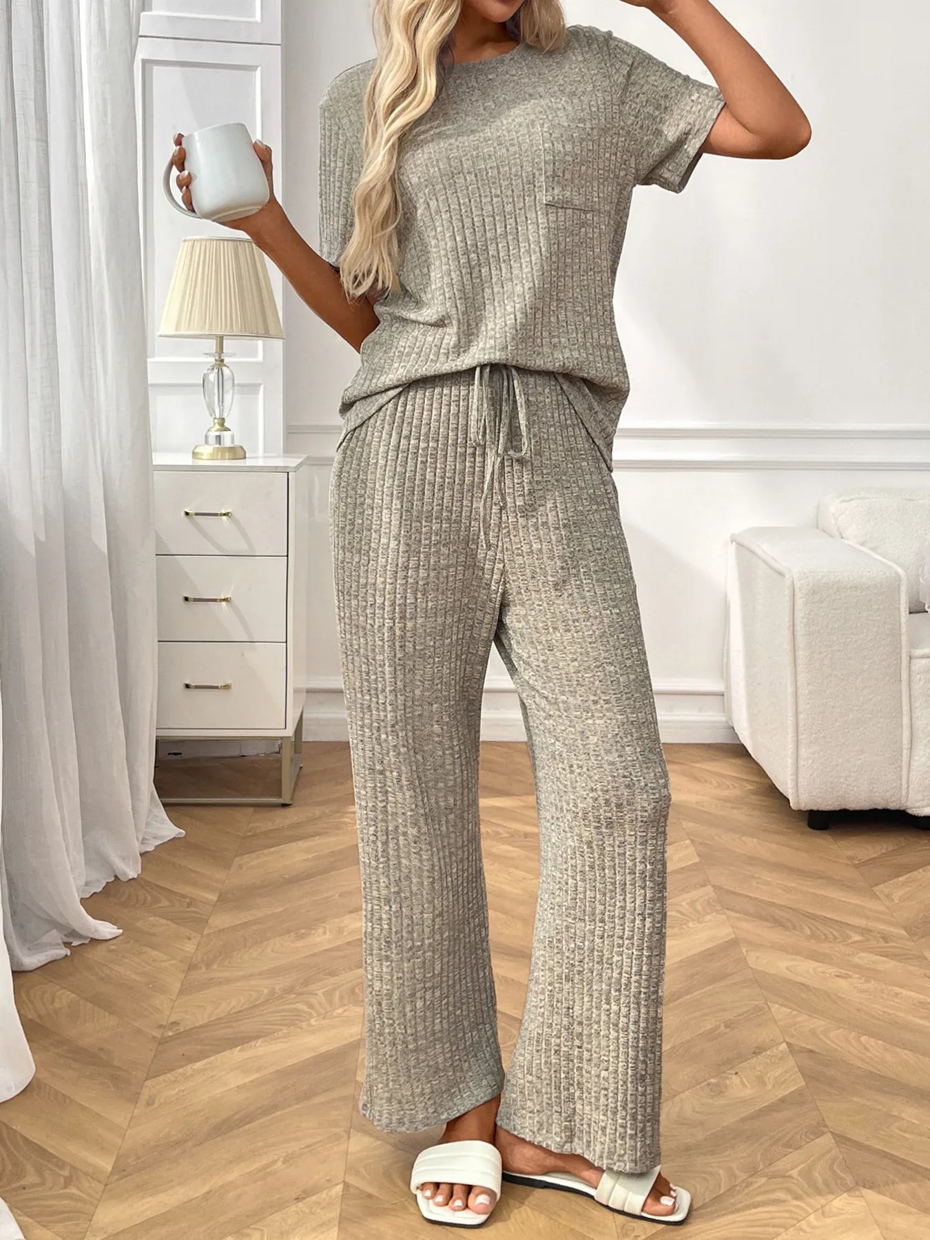 Woman wearing a grey textured pants and top set in a room with white furniture and decor.