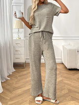 Woman wearing a grey textured pants and top set in a room with white furniture and decor.