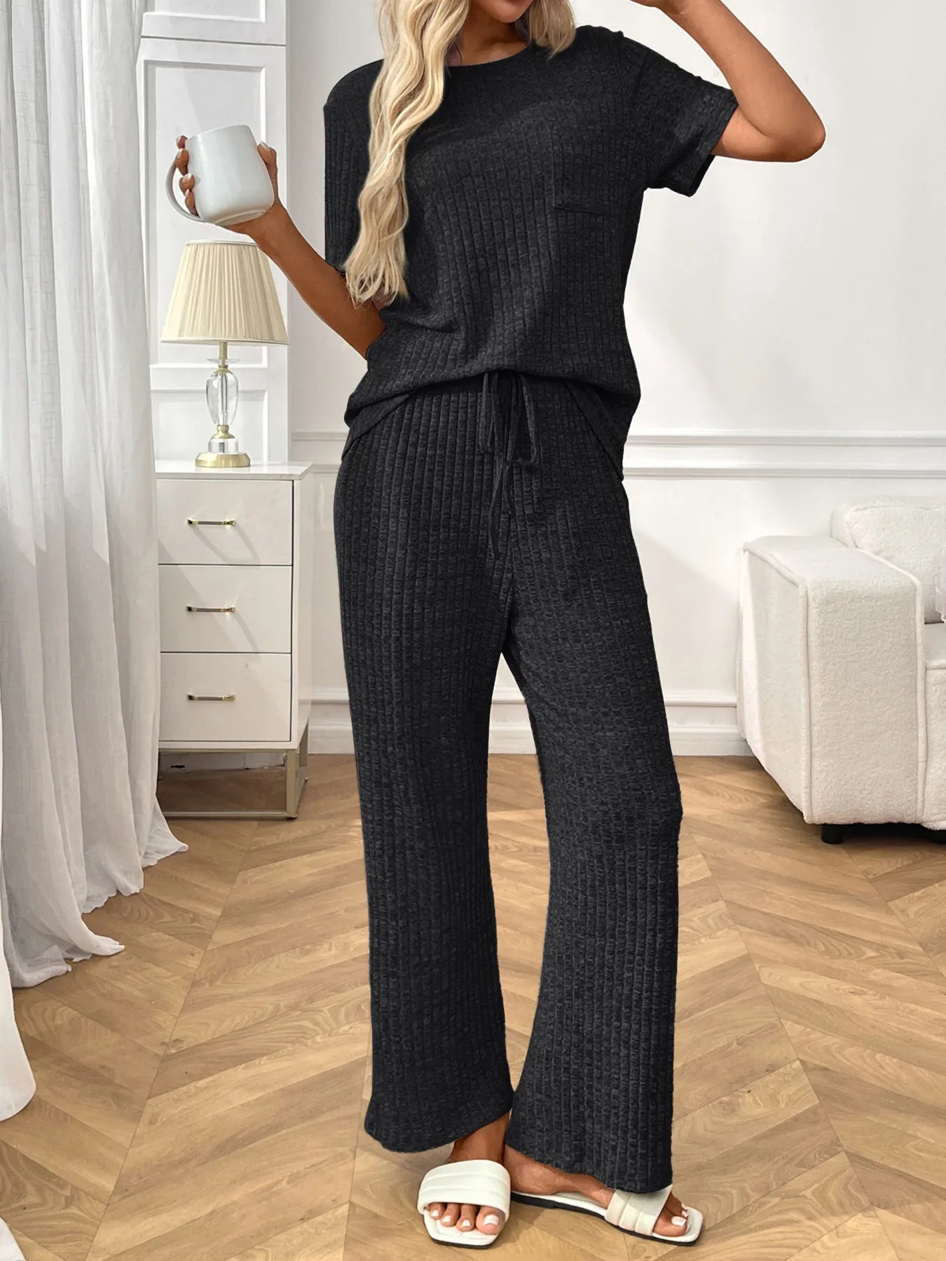 Woman wearing a black  textured pants and top set in a room with white furniture and wooden floor.