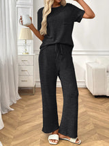 Woman wearing a black  textured pants and top set in a room with white furniture and wooden floor.
