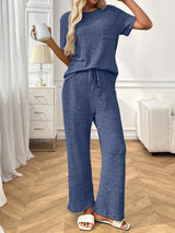 Woman in a blue  textured pants and top set  holding a mug in a room with white furniture and wooden floor.