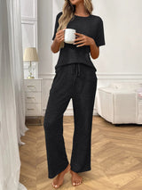 Woman in black textured pants and top set holding a white mug in a room with white furniture and decor.