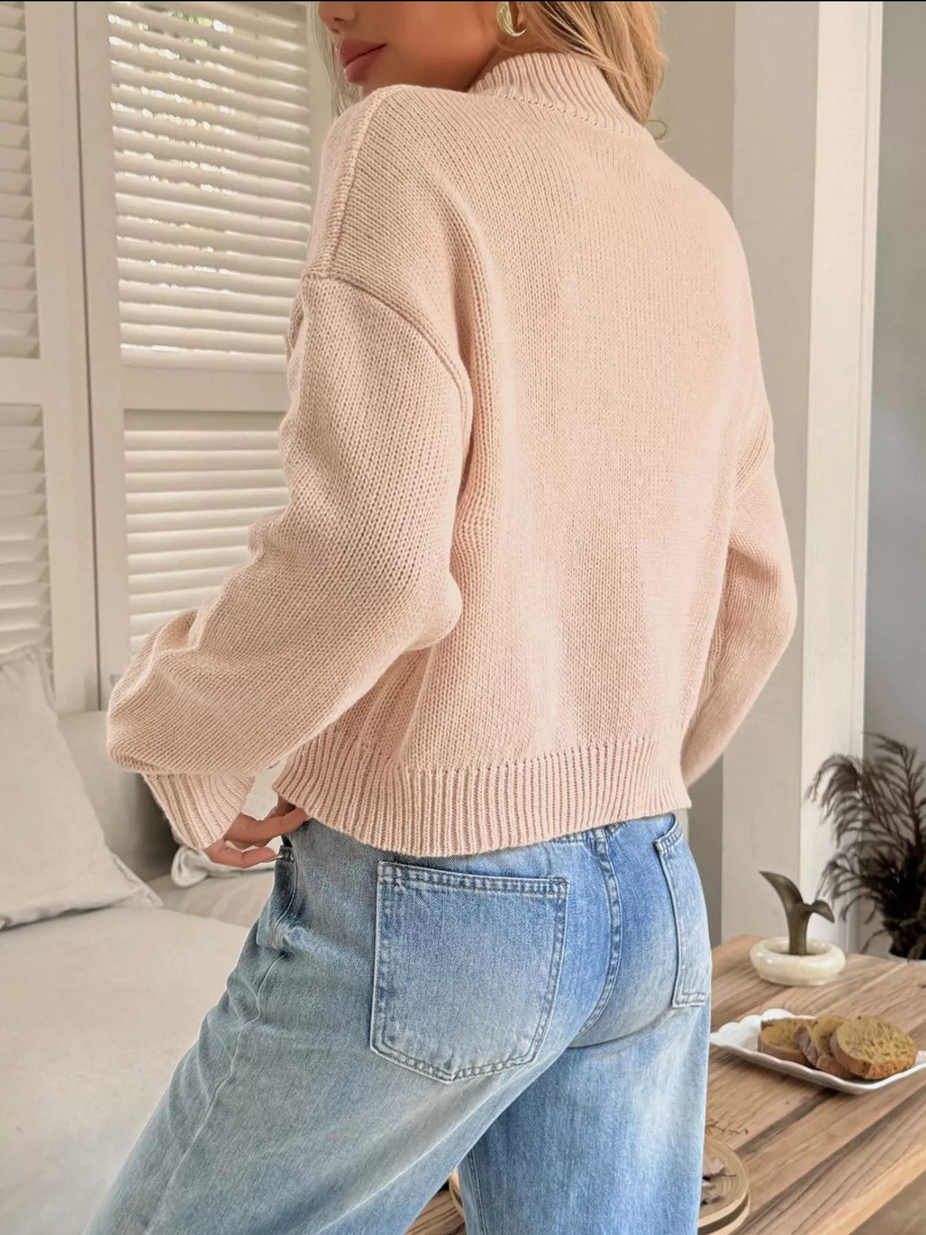 Person wearing a beige sweater and blue jeans in a room with a bed and window blinds.