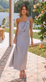 Woman in a light grey one-shoulder dress standing outdoors with greenery and a pool in the background.