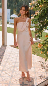 Woman in a beige one-shoulder dress standing outdoors near flowers and a building.