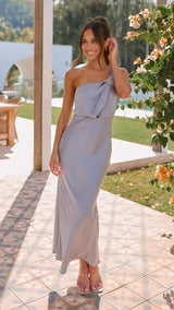 Woman in a gray one-shoulder dress standing outdoors near a garden.
