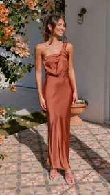 Woman in a rust-colored dress standing outdoors with flowers and a wall in the background