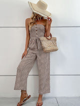 A woman wears The Janisa Jumpsuit in khaki, styled with a waist tie and wide-leg pants, standing confidently outside a white house.