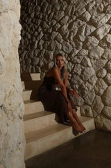 Woman wearing a brown halter neck maxi dress sitting on stone steps against a textured stone wall