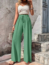 Woman wearing a white lace top and green wide-leg pants standing against a stone wall.