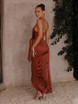 Woman in a rust-colored dress standing against a textured wall with greenery.