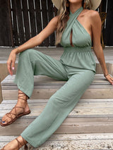 Model sitting on wooden steps wearing The Sage Halter Neck Jumpsuit with crossover bodice and wide-leg pants.